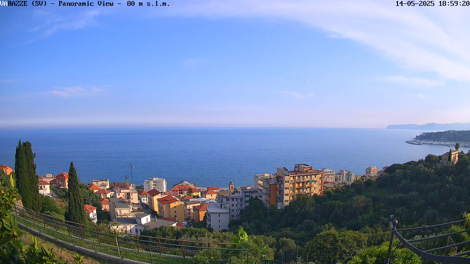 A coastal town with multi-story buildings is nestled on a hillside overlooking a calm blue sea under a clear, sunny sky.