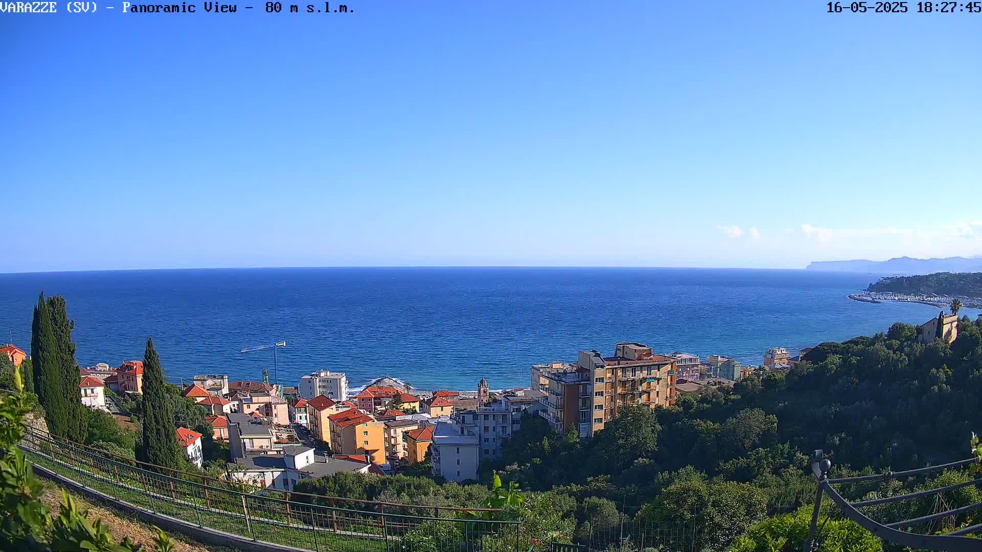 A panoramic view shows a coastal town nestled among green hills overlooking a calm, blue sea under a clear sky.