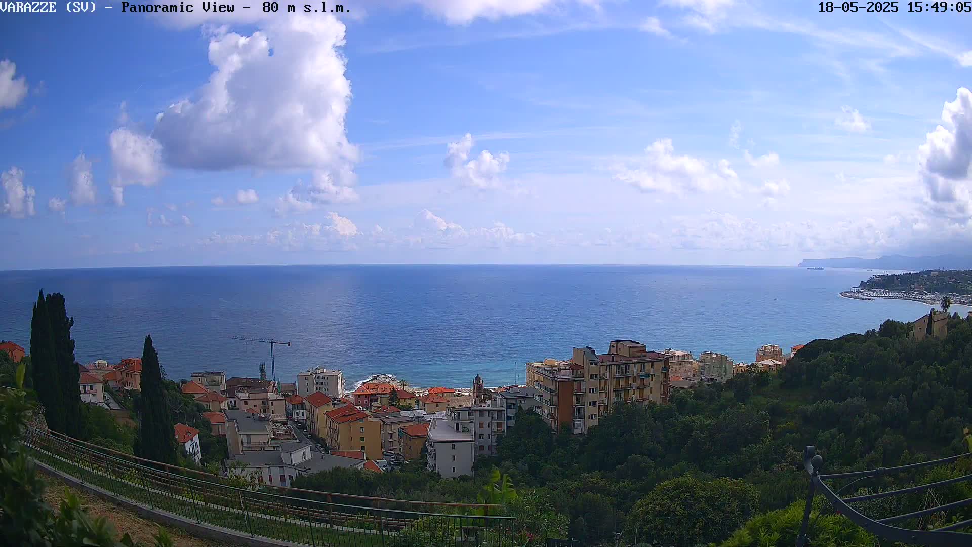 A partly cloudy day overlooks a coastal town situated on a hillside overlooking a calm, blue sea stretching to a distant shoreline.