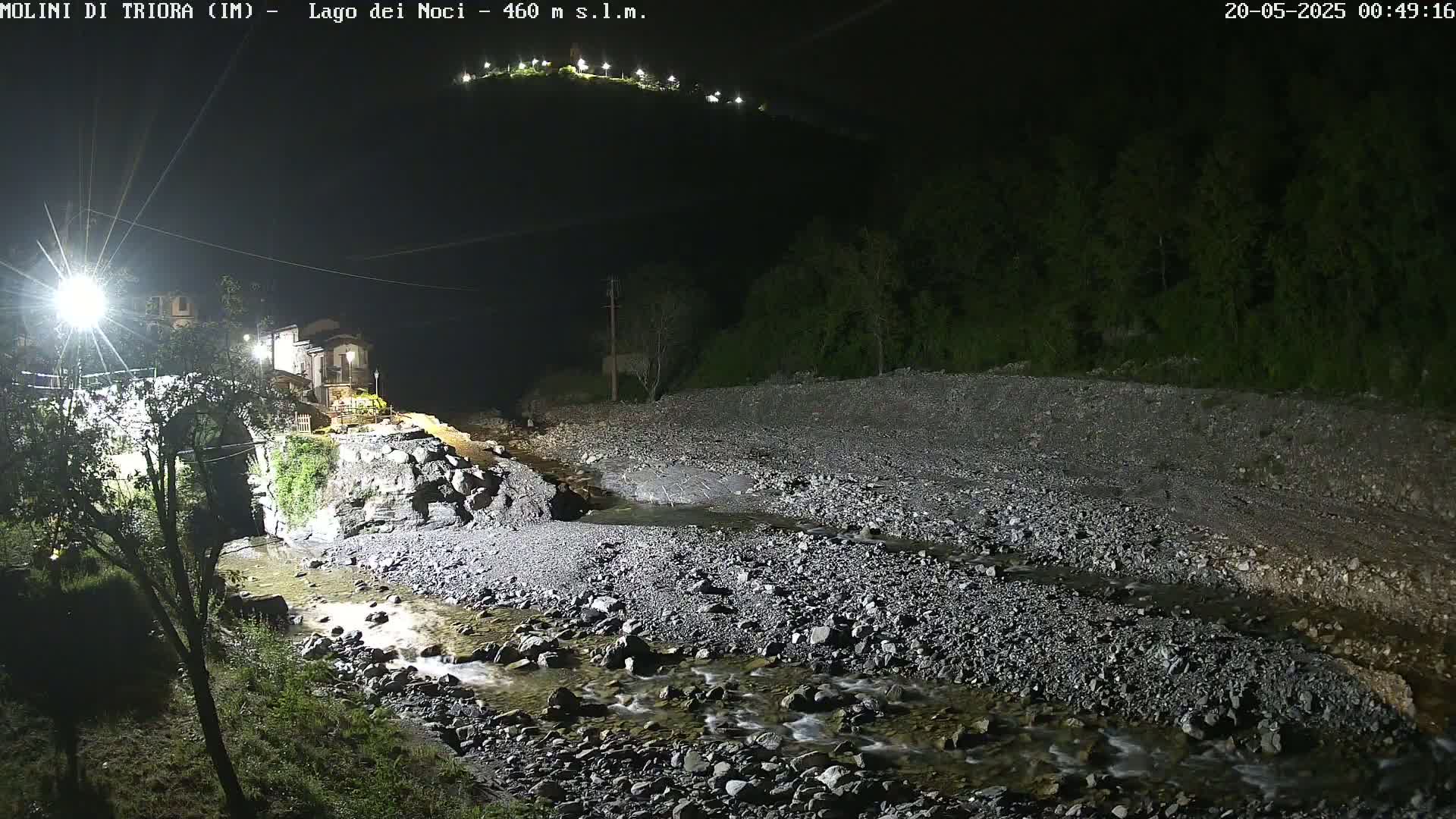 A rocky, shallow stream flows through a dark, nighttime landscape past a stone bridge and village illuminated by artificial lights, with a hilltop visible in the distance.