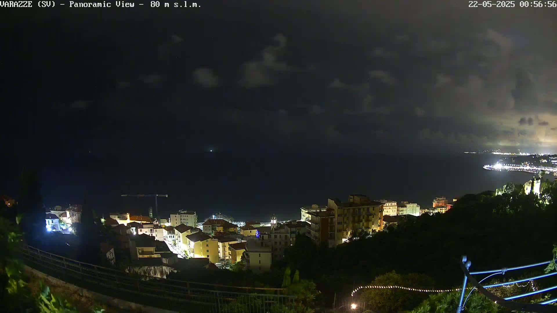 A nighttime panoramic view shows a coastal town with buildings lit up, situated on a hill overlooking a dark ocean under a partly cloudy sky.