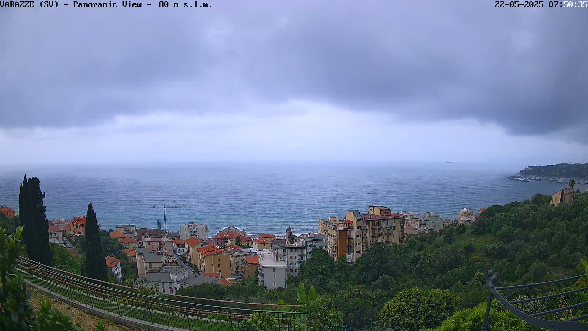 A panoramic view shows a coastal town nestled among green hills overlooking a calm sea under a cloudy sky.