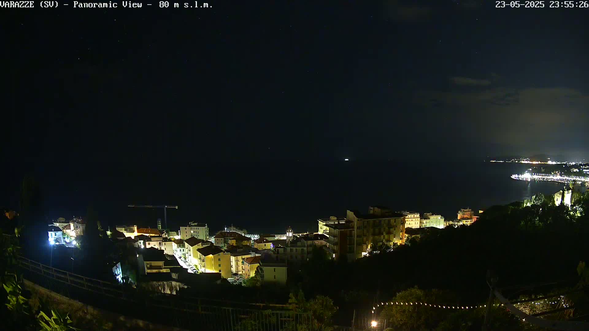 A nighttime panoramic view of a coastal town illuminated by lights, overlooking a dark sea under a starry sky.