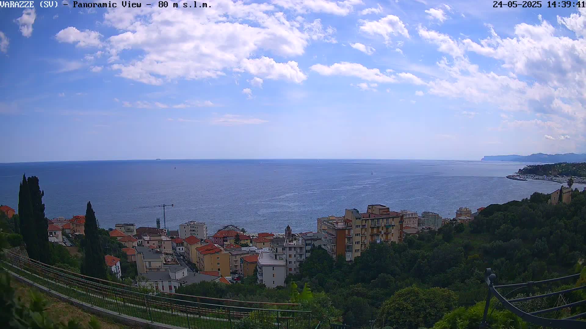 A partly cloudy day overlooking a coastal town with buildings and greenery, and a calm sea extending to a distant shoreline.