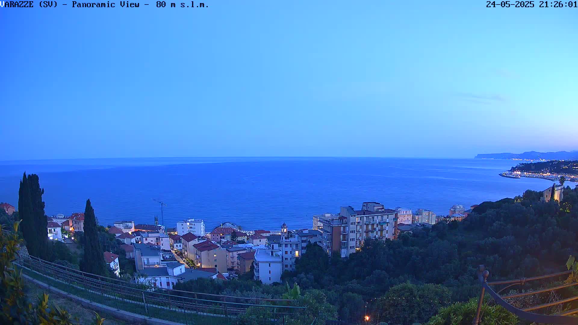 A coastal town at dusk is nestled on a hillside overlooking a calm, dark blue sea under a clear, twilight sky.