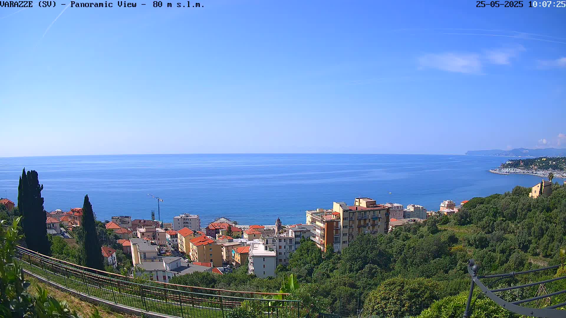 A coastal town with red-roofed buildings is nestled on a hillside overlooking a calm, blue sea under a mostly sunny sky.