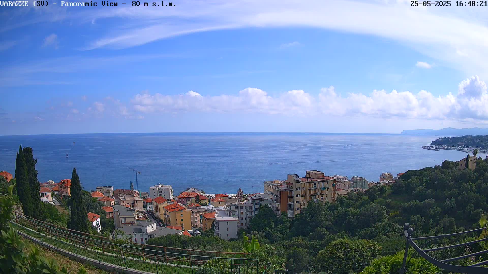 A panoramic view shows a coastal town nestled among green hills overlooking a calm blue sea under a partly cloudy sky.
