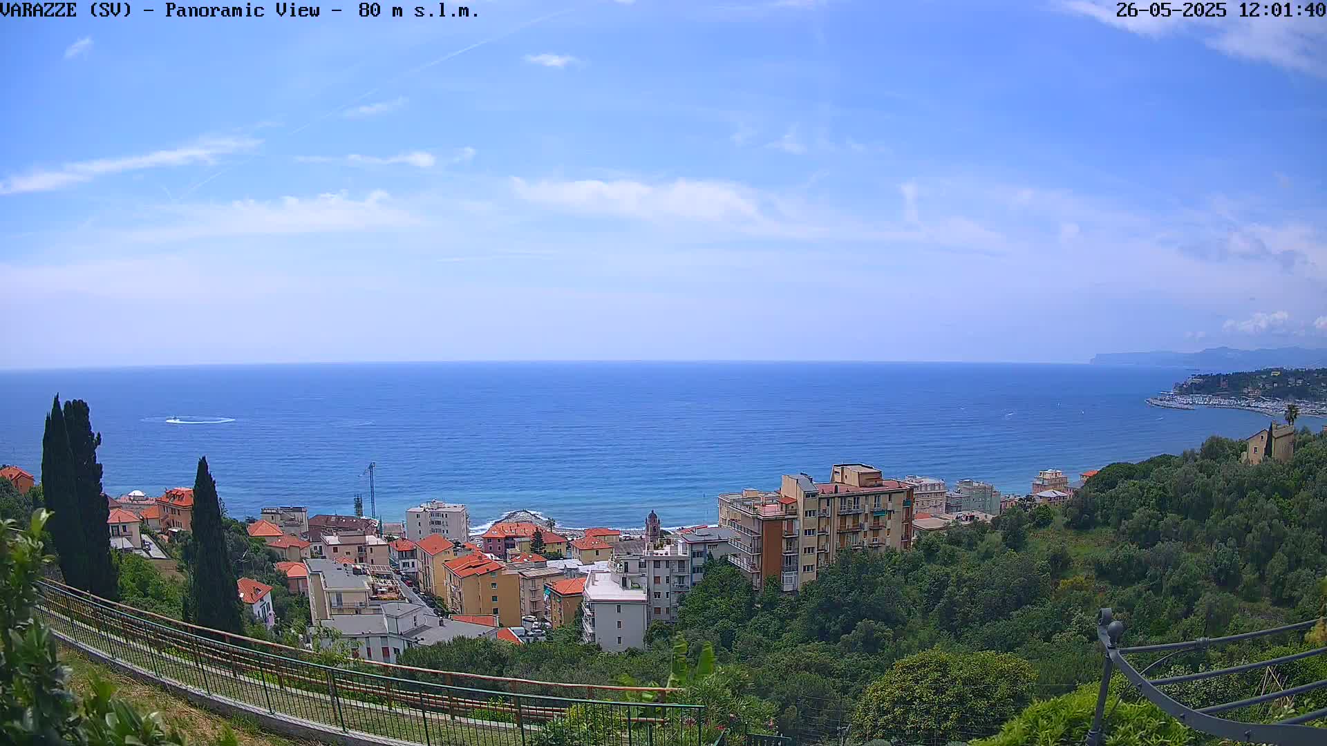 A panoramic view of a coastal town on a partly cloudy day, with a calm blue sea extending to a distant shoreline.