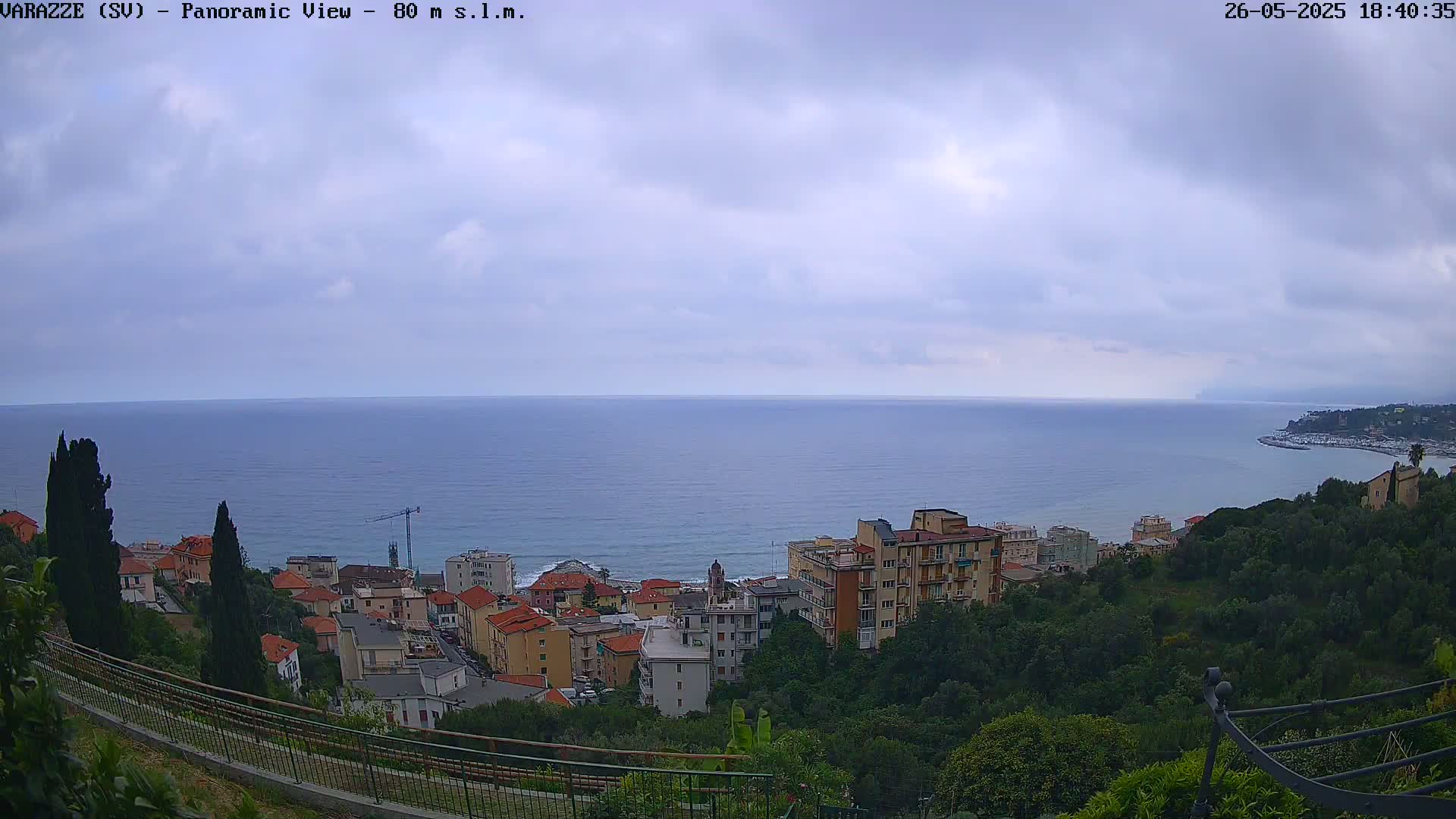A coastal town with multi-story buildings is nestled on a hillside overlooking a calm sea under a cloudy sky.