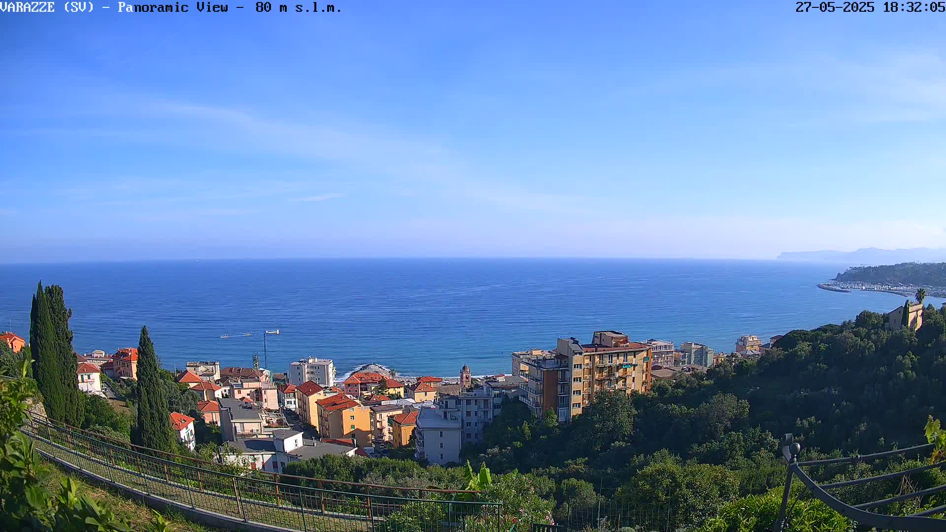 A panoramic view shows a coastal town nestled on a hillside overlooking a calm blue sea under a clear sky.