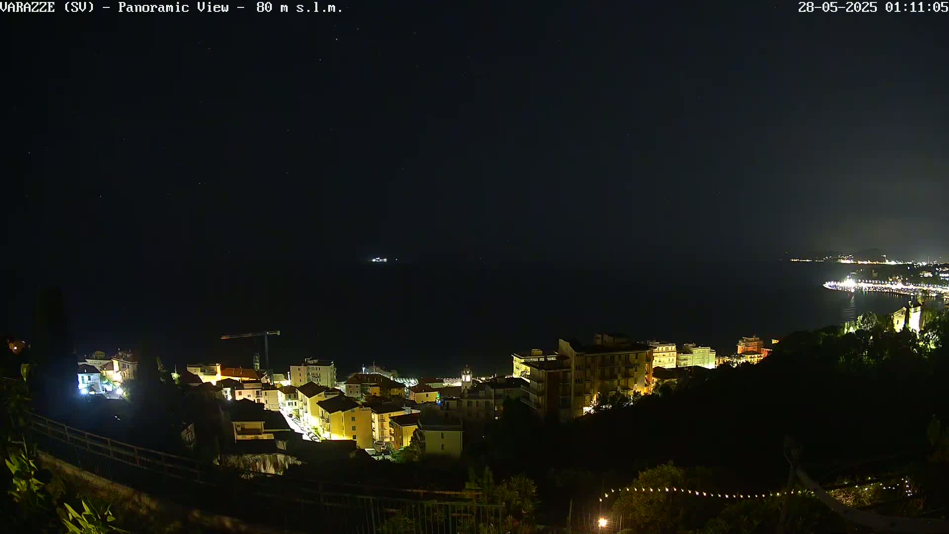 A nighttime, panoramic view shows a coastal town with illuminated buildings overlooking a dark ocean under a clear, starry sky.