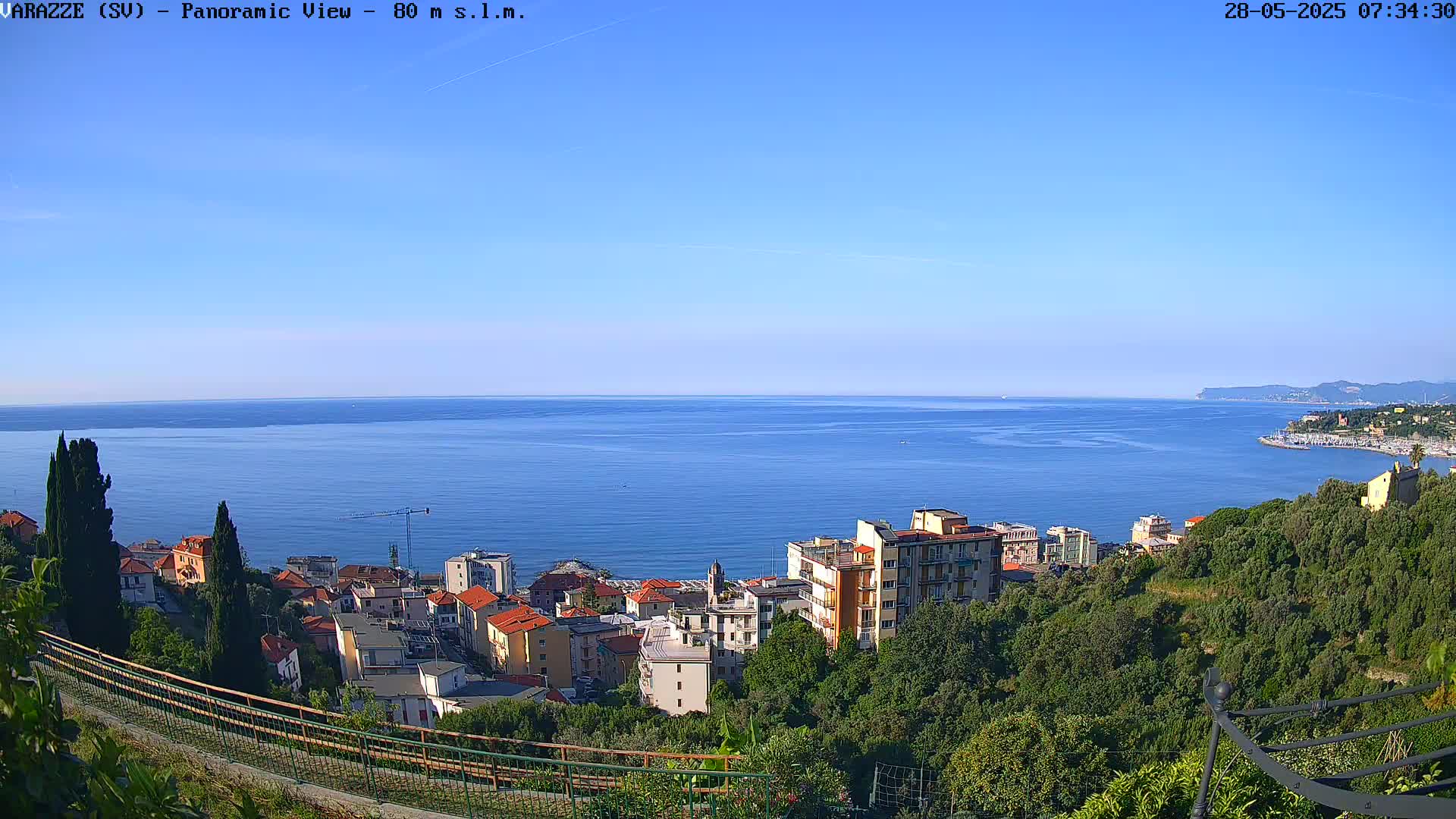 A panoramic view of a coastal town nestled amongst lush green hills overlooking a calm blue sea under a clear blue sky.