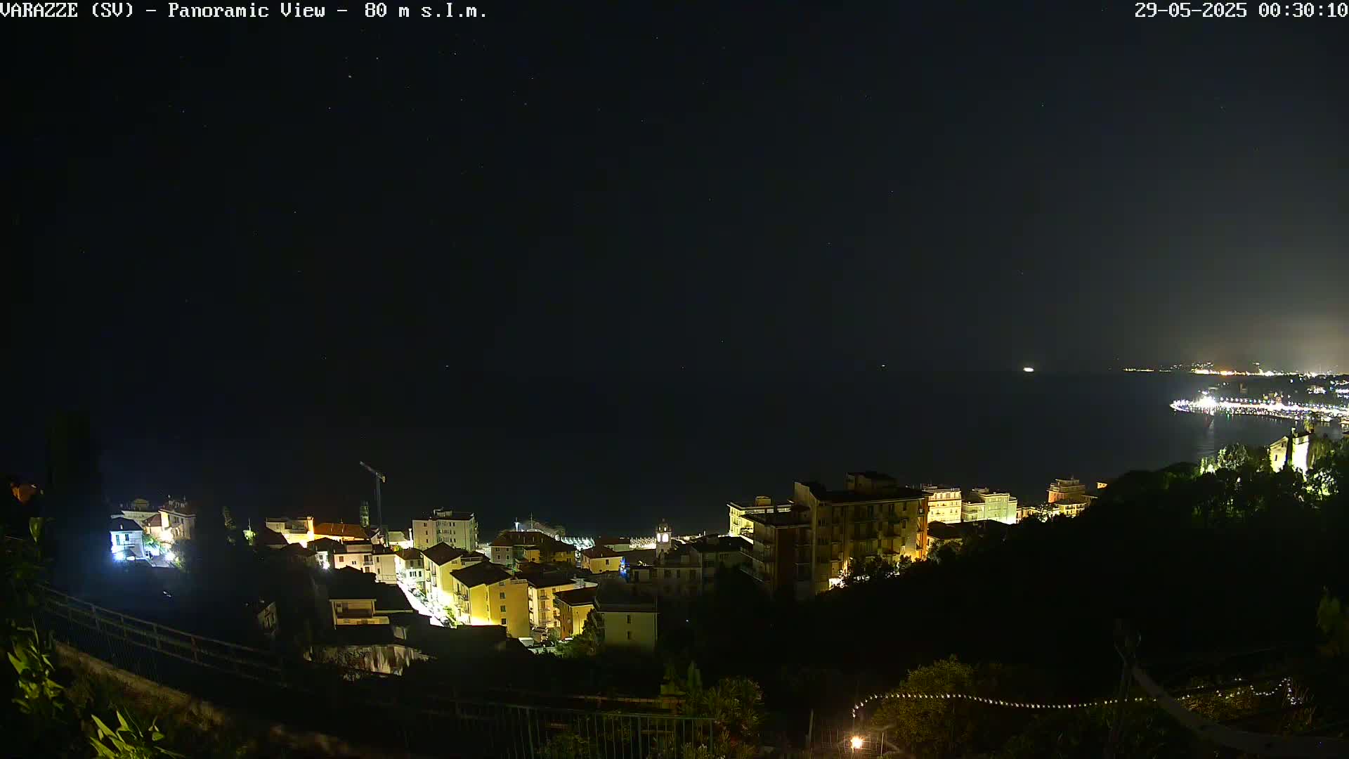 A nighttime panoramic view of a coastal town illuminated by lights, with a dark sea and starry sky in the background.