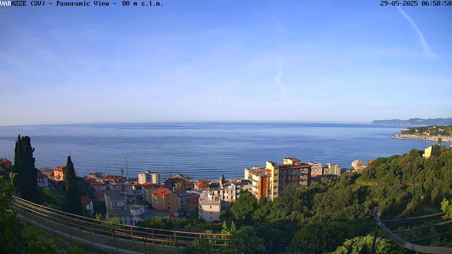 A panoramic view of a coastal town nestled among green hills overlooking a calm, blue sea under a clear sky.