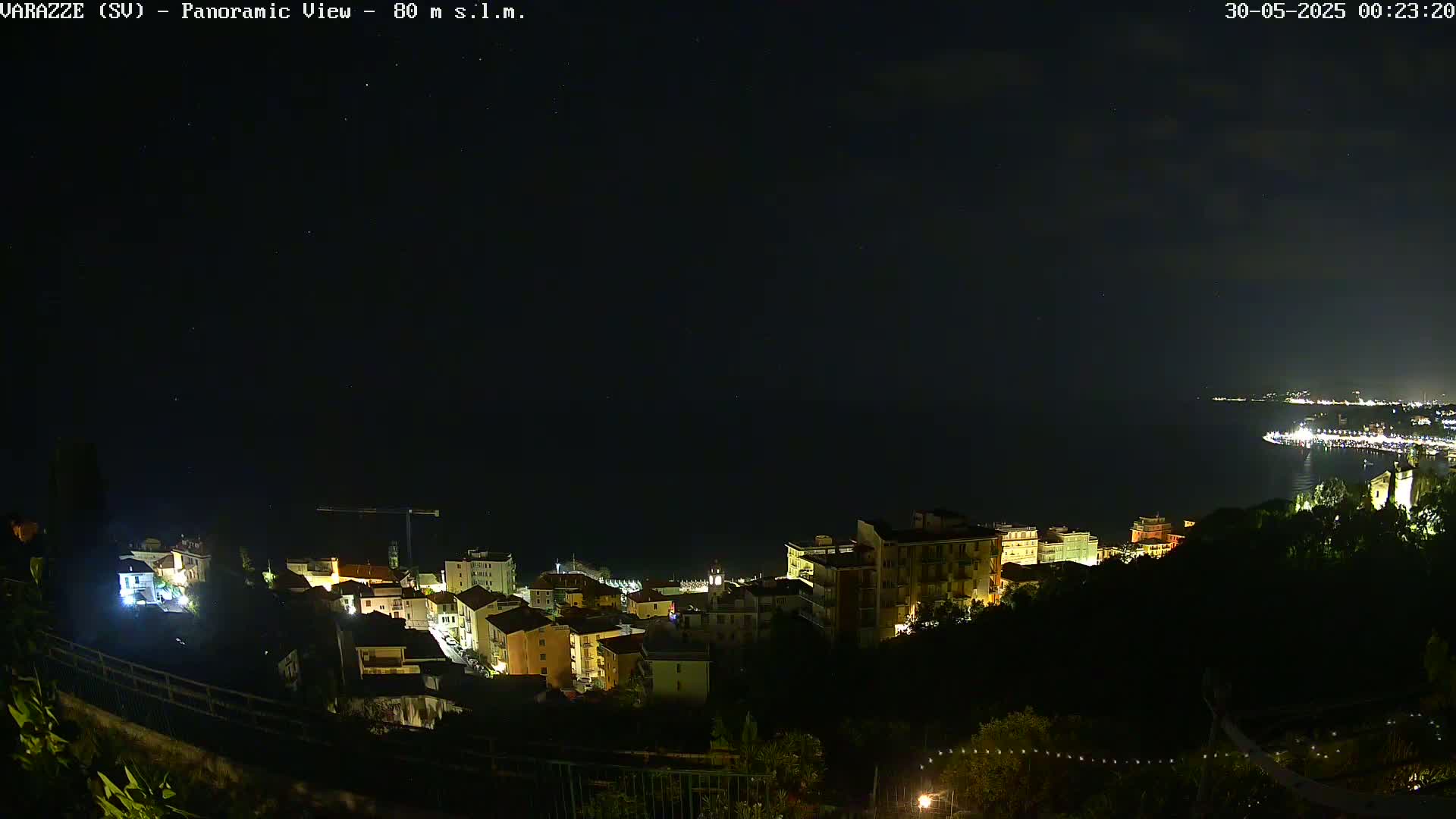 A nighttime panoramic view shows a coastal town with illuminated buildings nestled on a hillside overlooking a dark sea under a clear, starry sky.