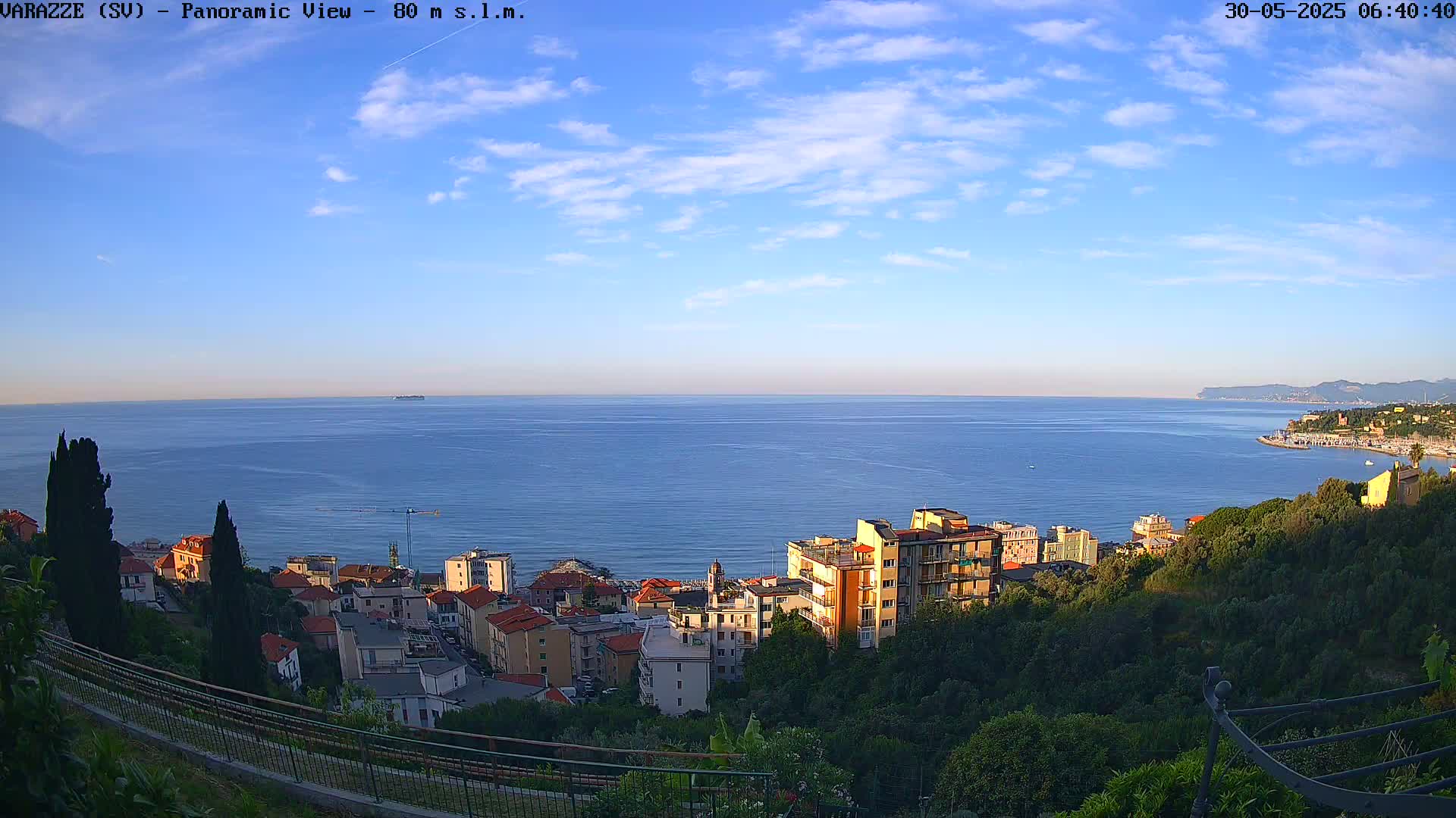 A coastal town nestled on a hillside overlooks a calm, blue sea under a mostly clear, bright sky.
