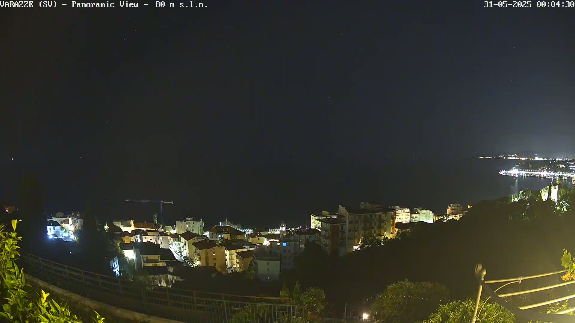 A nighttime, panoramic view of a coastal town shows buildings illuminated against a dark sky speckled with stars, and a calm sea.