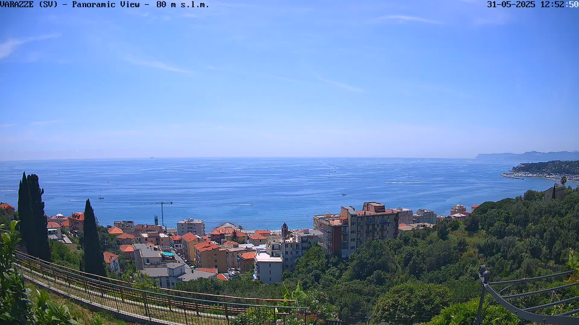A coastal town nestled among lush green hills overlooks a calm, blue sea under a clear, sunny sky.