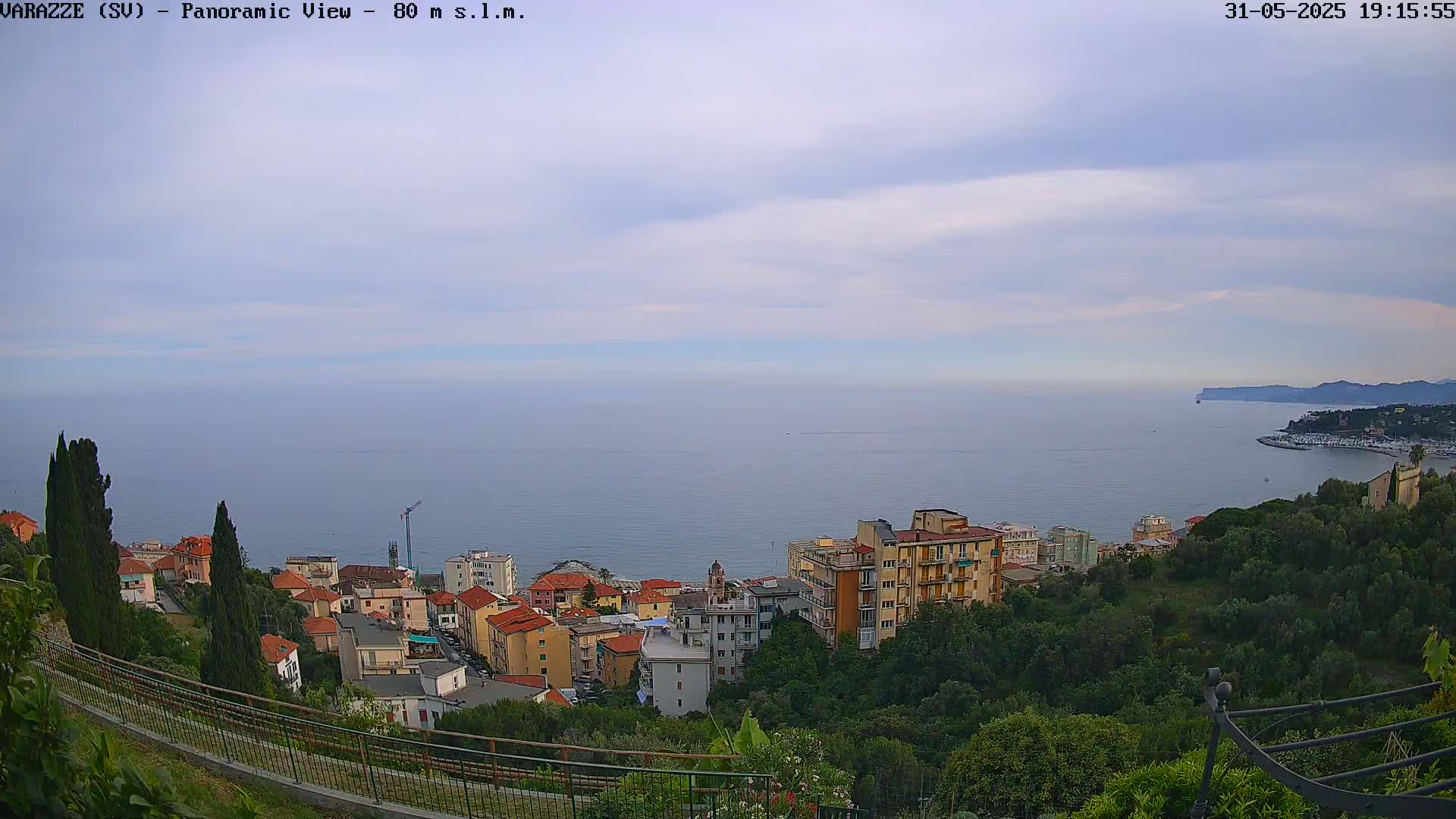 A coastal town with multi-story buildings nestled amongst green vegetation overlooks a calm sea under a partly cloudy sky.