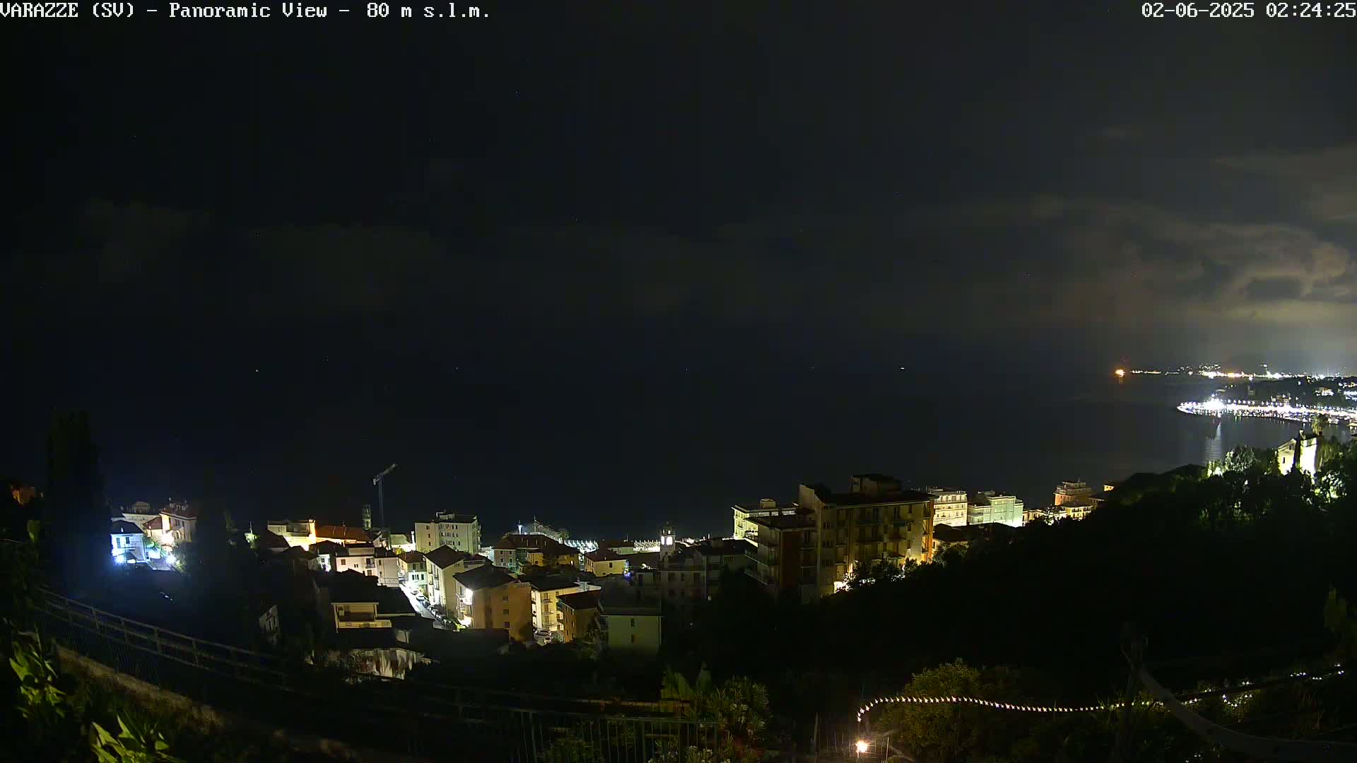 A nighttime panoramic view of a coastal town shows buildings lit up against a dark sky with some clouds and a calm sea.
