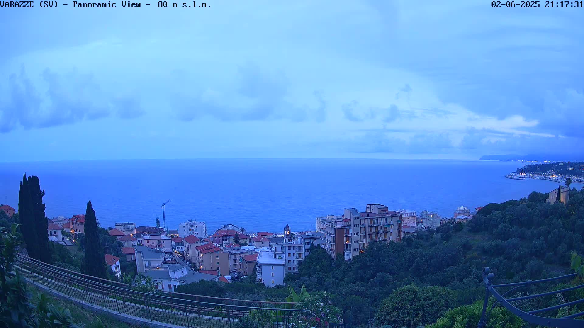 A coastal town sits on a hillside overlooking a calm, twilight sea under a partly cloudy sky.