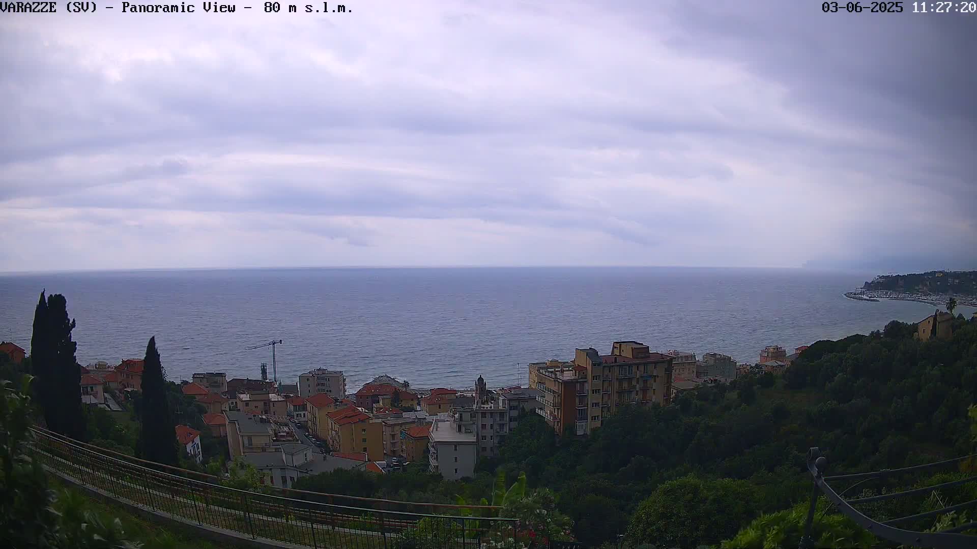 A coastal town with multi-story buildings is nestled among green hills overlooking a calm sea under a cloudy sky.