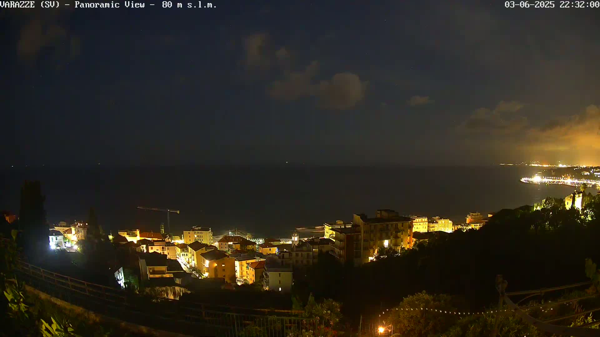 A nighttime panoramic view of a coastal town illuminated with lights, under a partly cloudy sky, overlooking a dark sea.