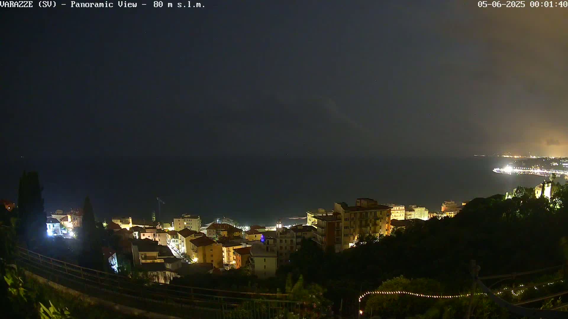 A nighttime panoramic view shows a coastal town illuminated, overlooking a dark sea under an overcast sky.