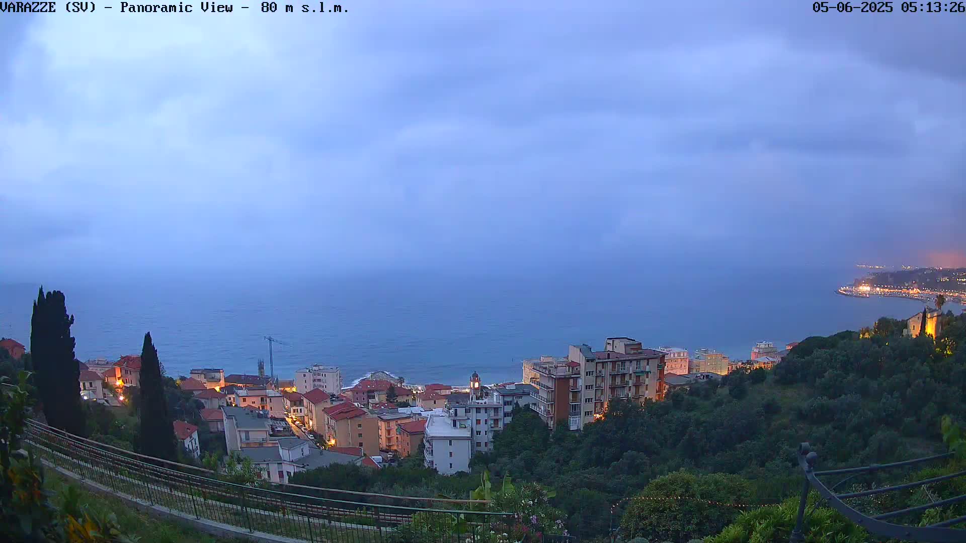 A coastal town with buildings illuminated at twilight under a cloudy sky overlooks a calm sea.