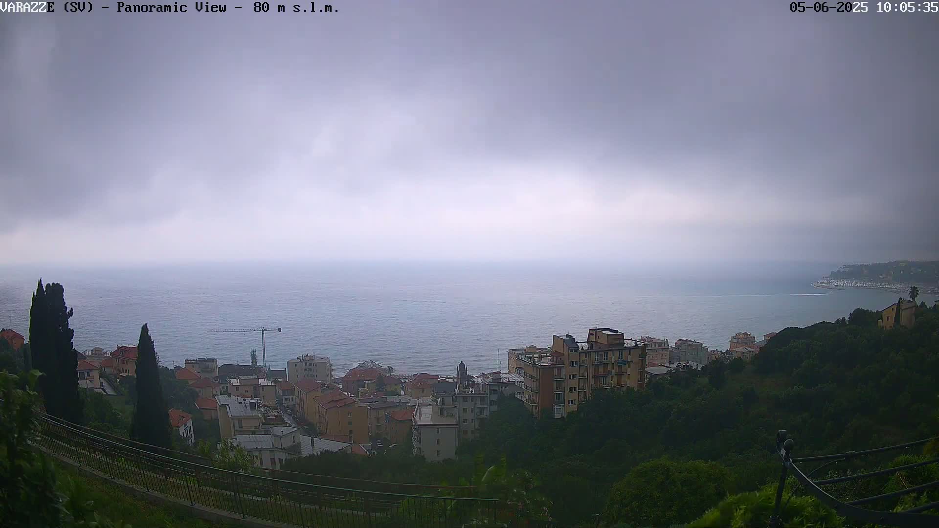 A panoramic view of a coastal town under an overcast sky shows a calm sea extending to the horizon, with buildings nestled on a hillside overlooking the water.