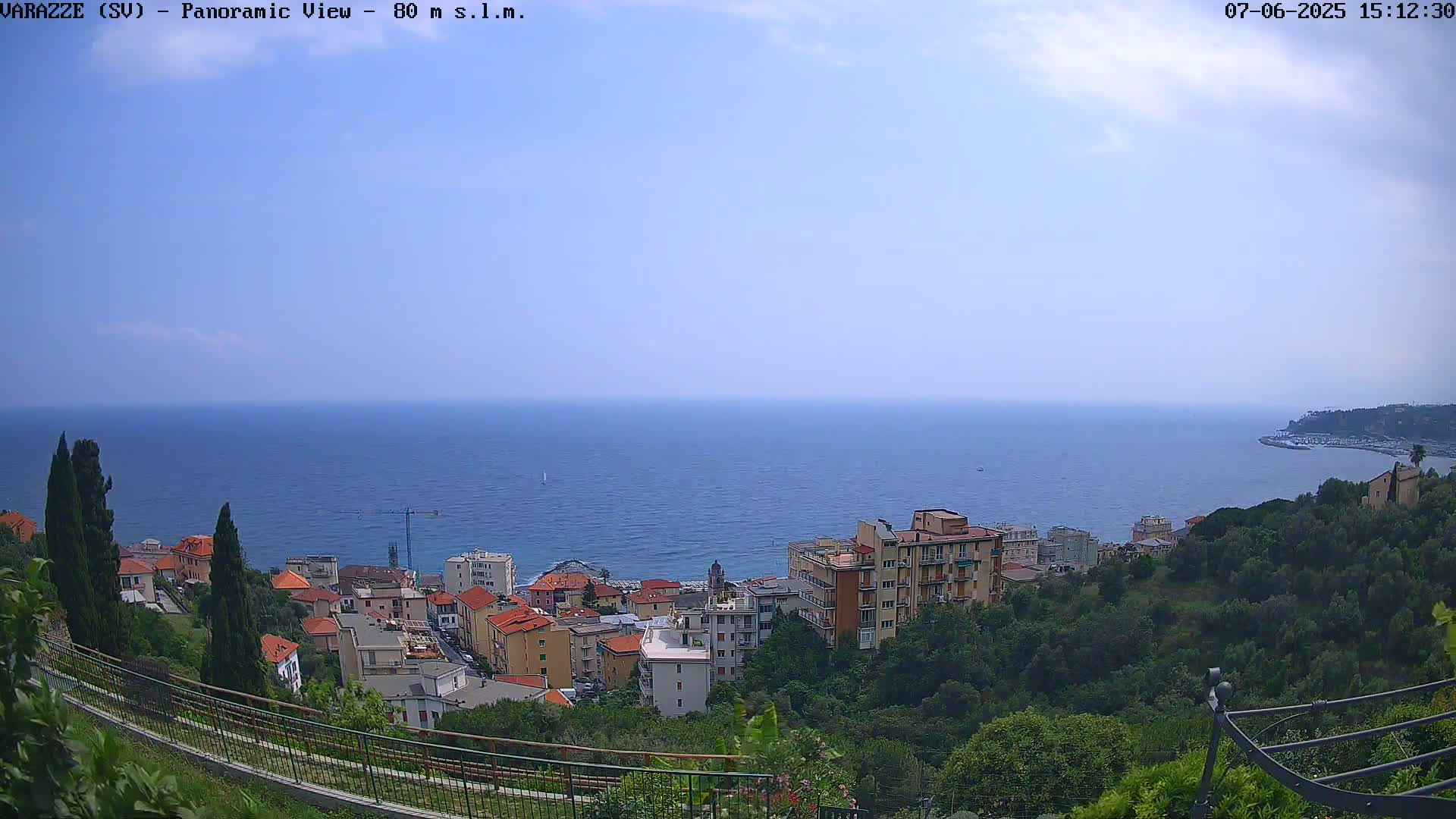 A coastal town nestled on a hillside overlooks a calm, blue sea under a mostly clear sky.
