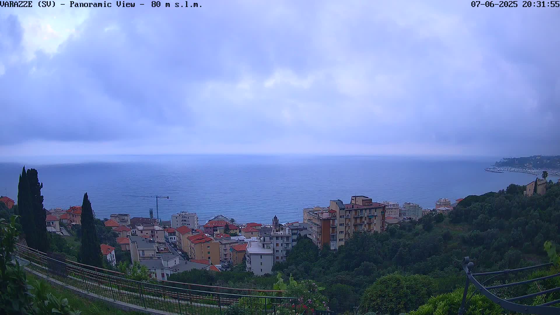 A panoramic view shows a coastal town nestled among green hills overlooking a calm sea under a cloudy sky.