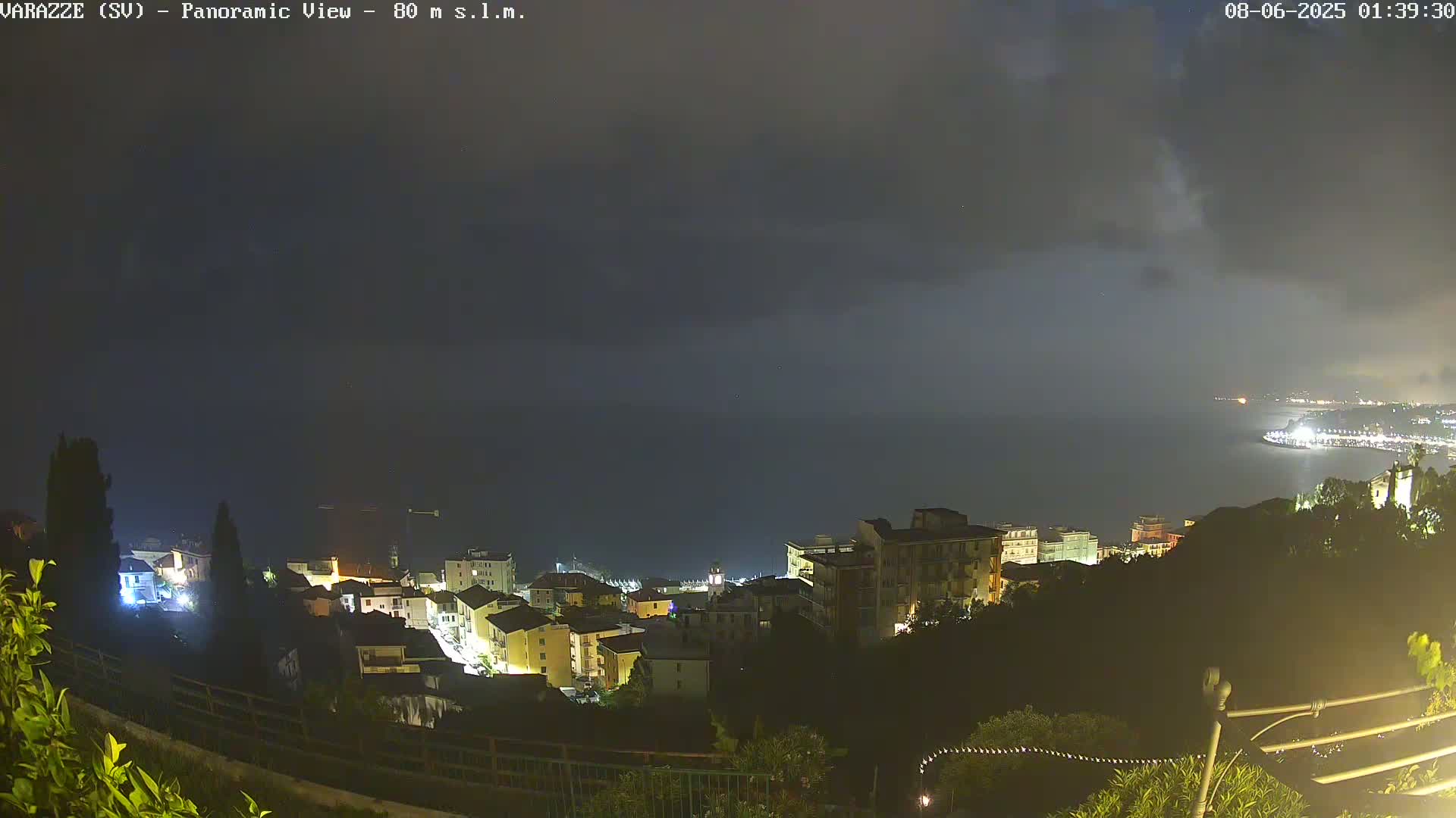 A nighttime, overcast view of a coastal town, with its lights illuminating buildings on a hillside overlooking a dark sea.
