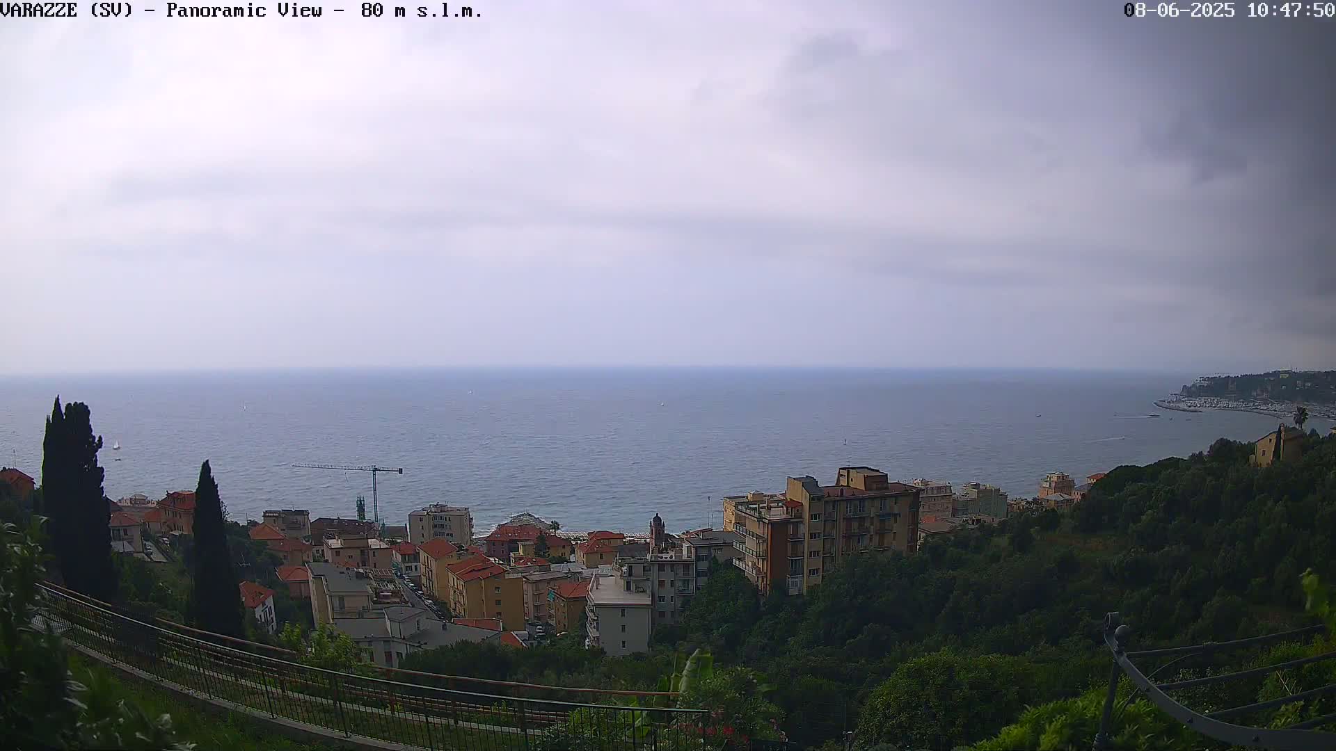 A seaside town with buildings and trees on a hillside overlooks a calm sea under a cloudy sky.