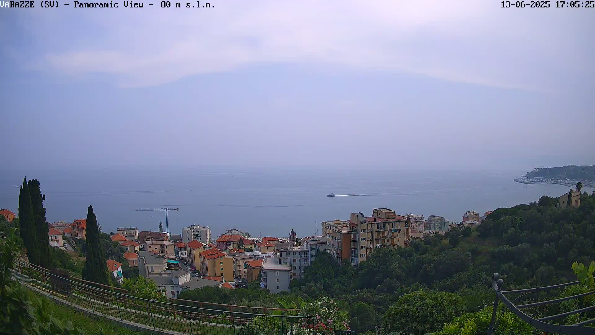 A panoramic view of a coastal town on a hazy day shows buildings nestled among green hills overlooking a calm sea.