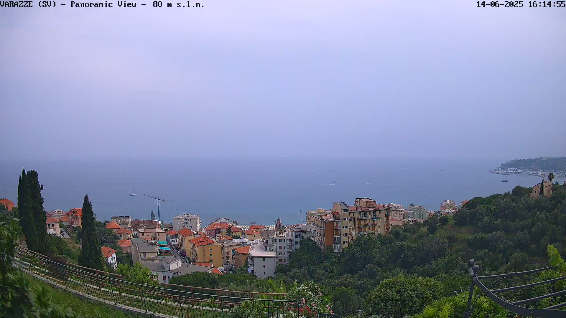 A hazy view overlooking a coastal town with multi-story buildings, green hillsides, and a calm sea in the distance.