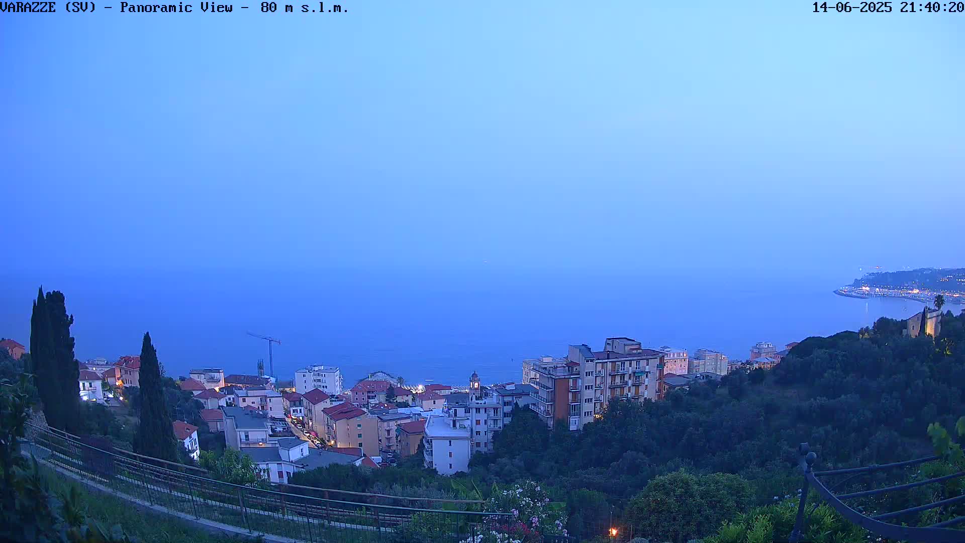 A hazy view overlooking a coastal town with multi-story buildings, green hillsides, and a calm sea in the distance.