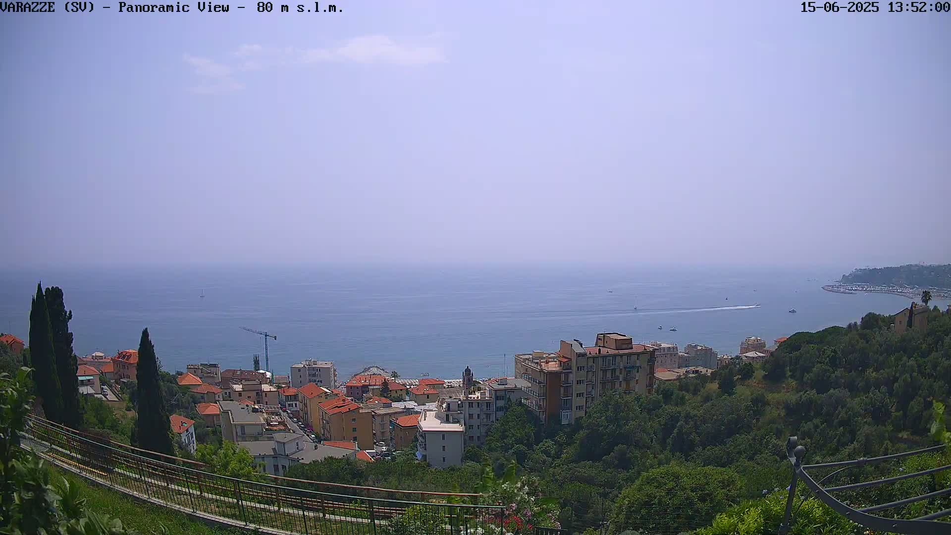 A panoramic view shows a coastal town nestled on a hillside overlooking a calm, hazy ocean on a clear day.