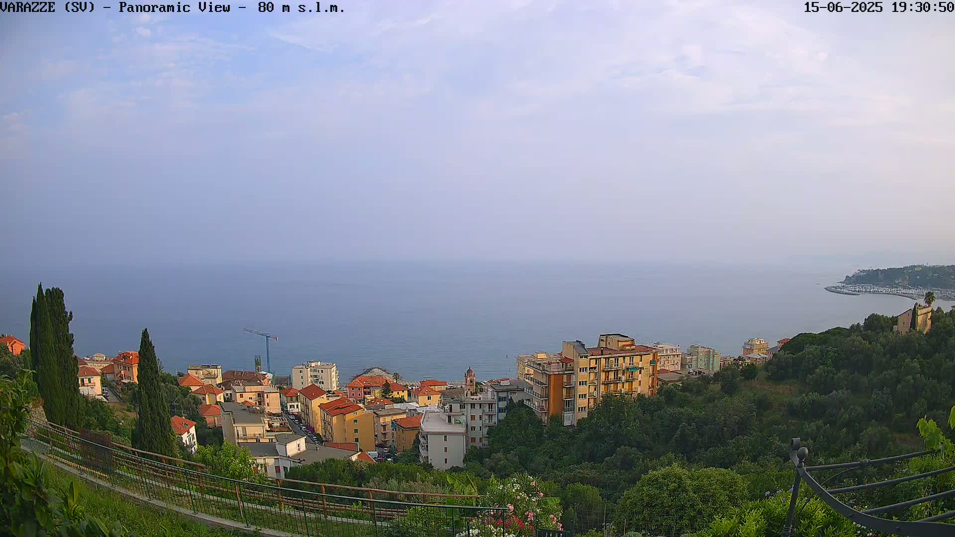 A panoramic view shows a coastal town nestled on a hillside overlooking a calm sea under a hazy sky.