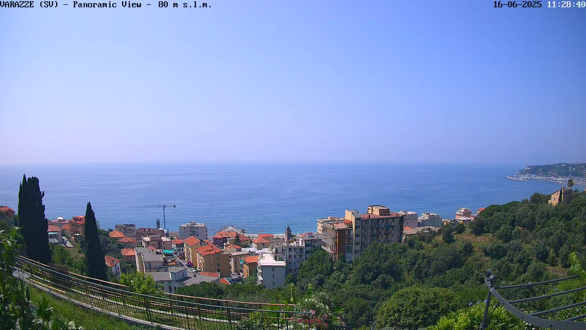 A panoramic view of a coastal town nestled amongst lush green hills overlooking a calm blue sea under a clear sky.