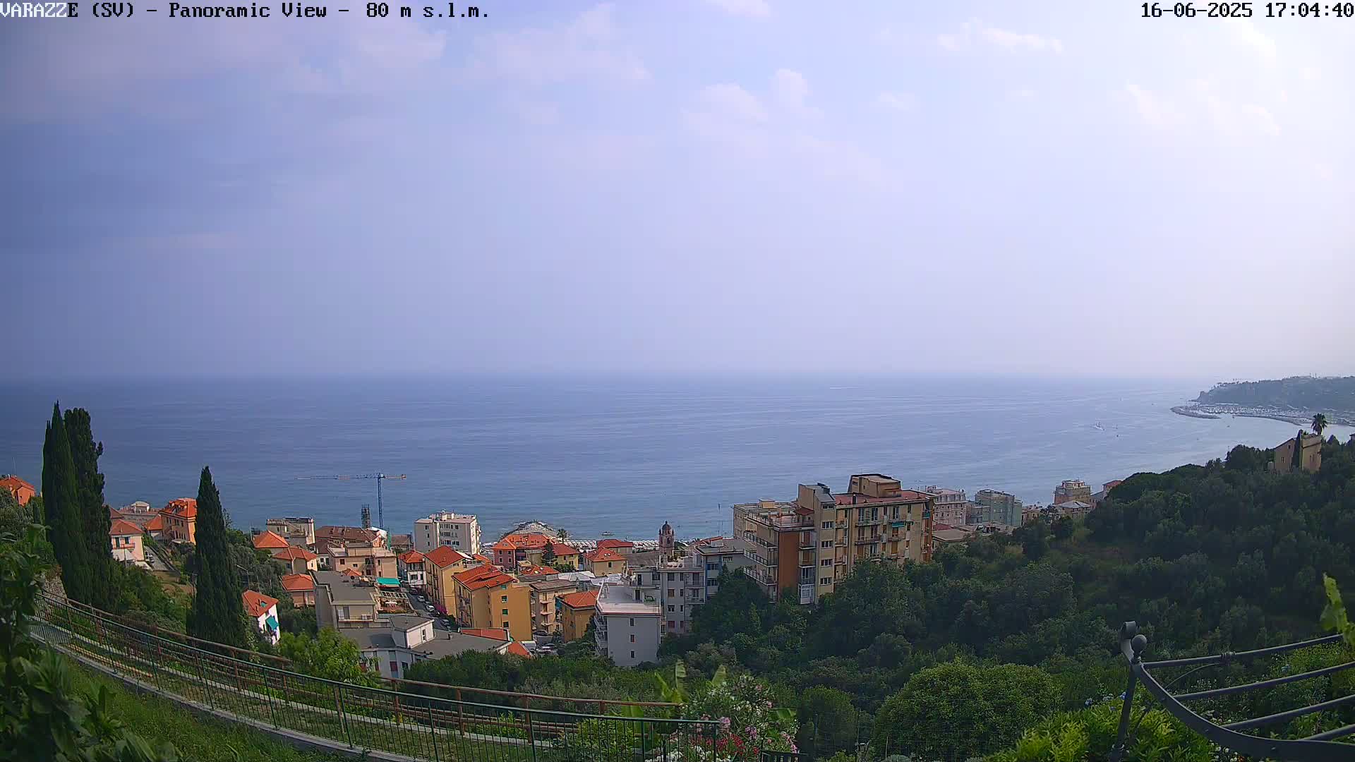 A panoramic view shows a coastal town nestled on a hillside overlooking a calm sea under a mostly clear sky with a few scattered clouds.