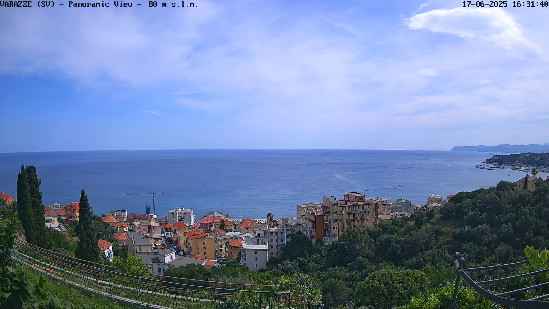 A coastal town with red-roofed buildings is nestled on a hillside overlooking a calm, blue sea under a mostly clear sky.
