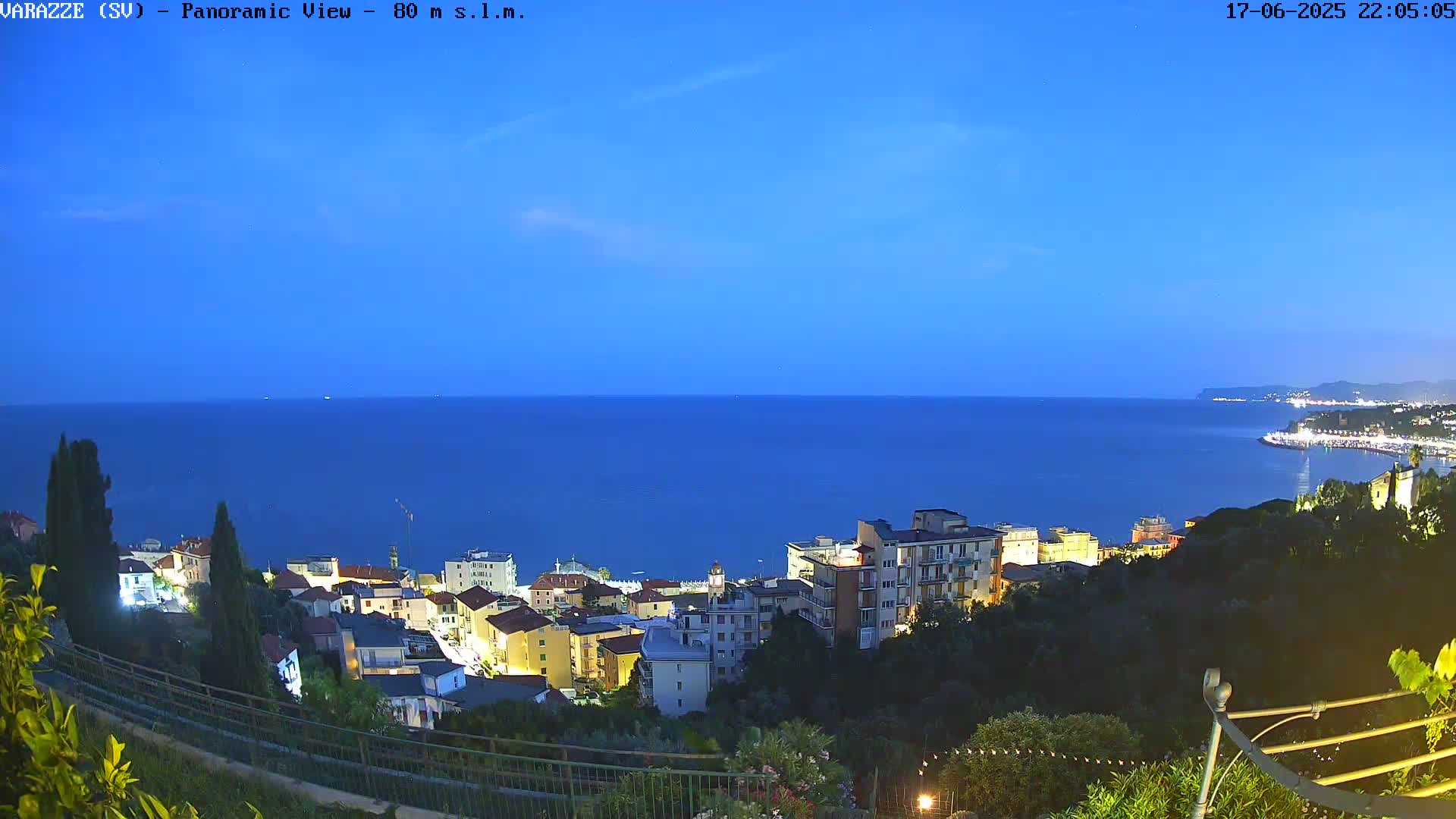 A coastal town at night, illuminated by streetlights, overlooks a calm, dark-blue sea under a twilight sky.