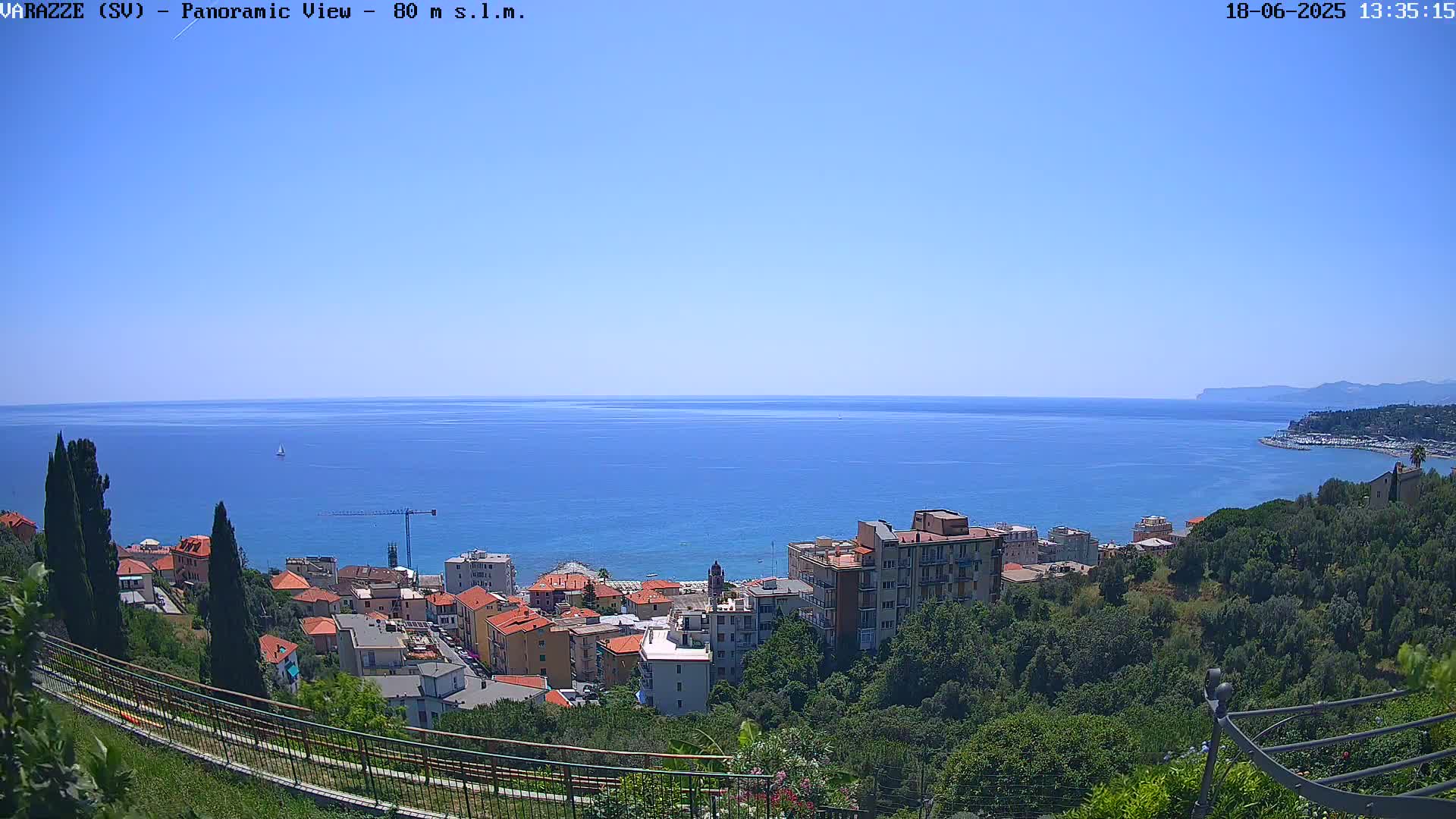 A coastal town nestled amongst green hills overlooks a calm, blue sea under a clear sky.