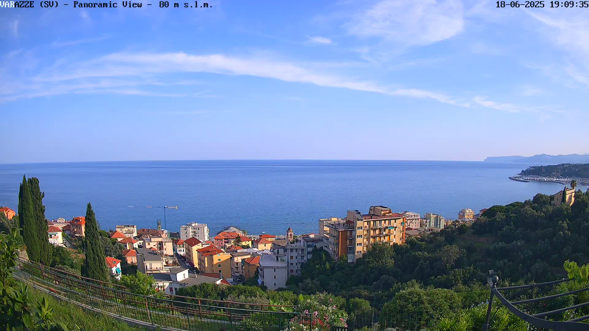 A coastal town with colorful buildings is nestled on a hillside overlooking a calm, blue sea under a mostly clear, sunny sky.