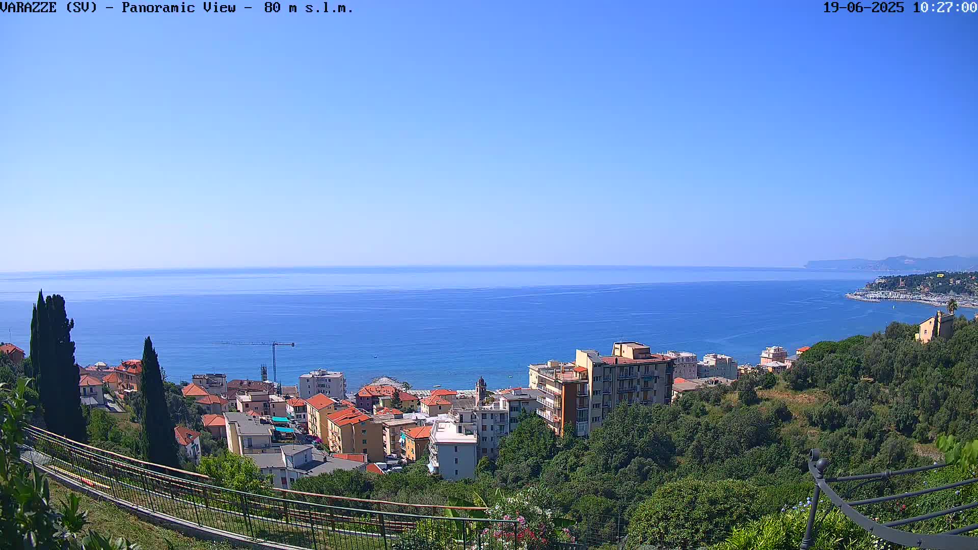 A panoramic view of a coastal town nestled among green hills overlooking a calm blue sea under a clear sky.