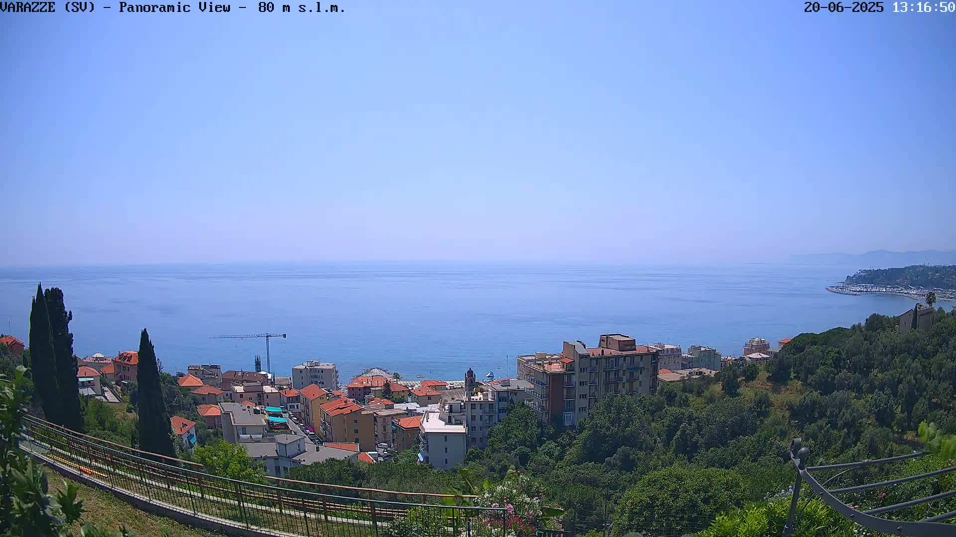 A coastal town nestled among green hills overlooks a calm, blue sea under a clear sky.