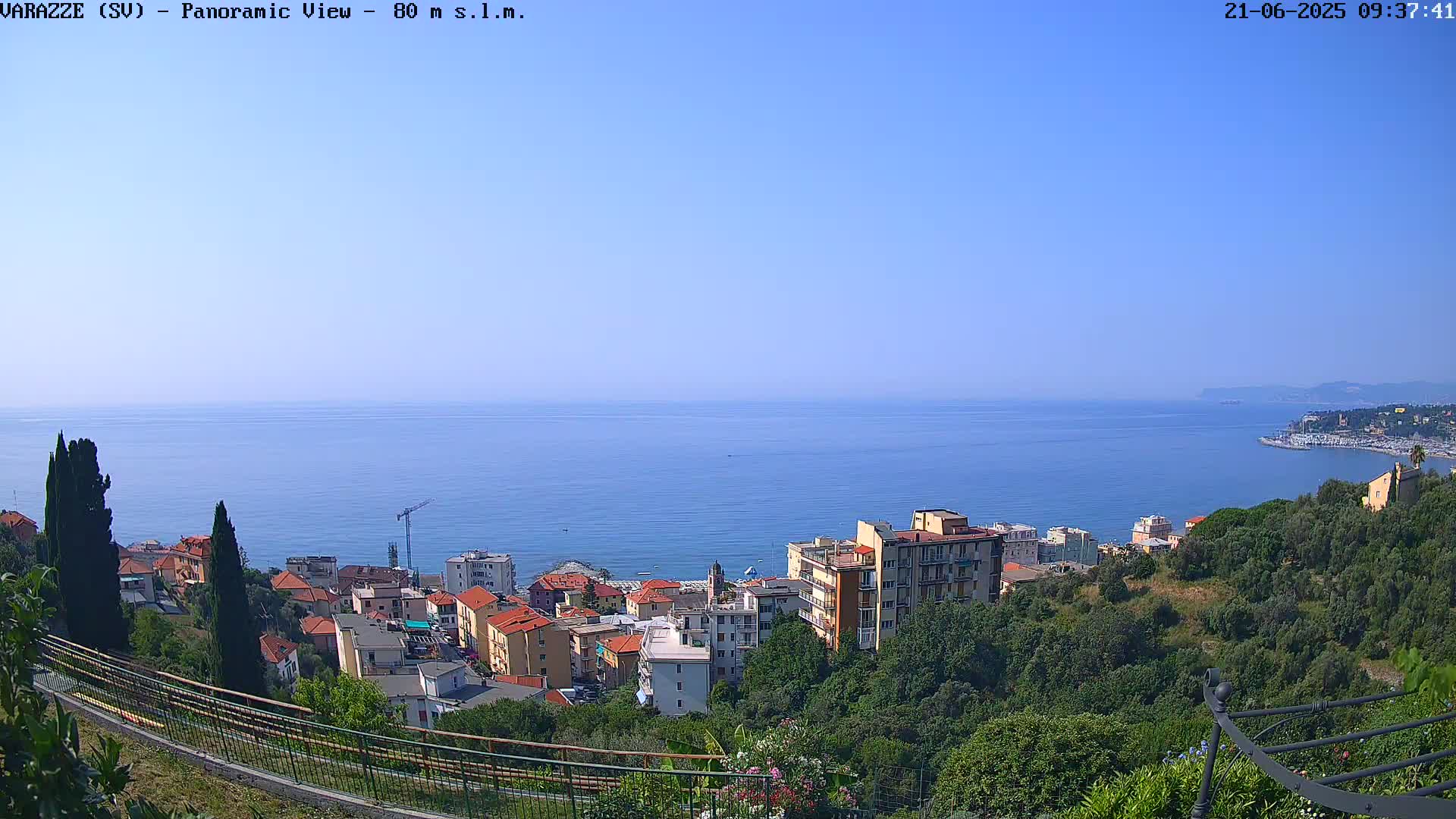 A panoramic view of a coastal town nestled on a hillside overlooking a calm blue sea under a clear sky.