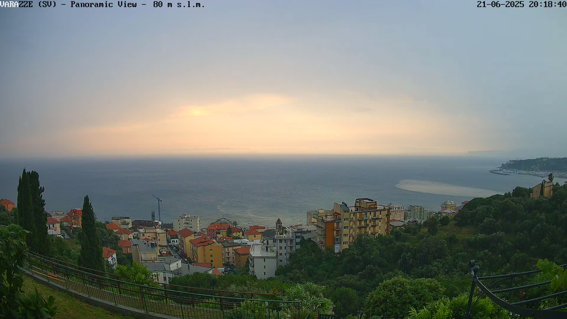 A coastal town with pastel-colored buildings is nestled on a hillside overlooking a calm sea under a partly cloudy, dusky sky.
