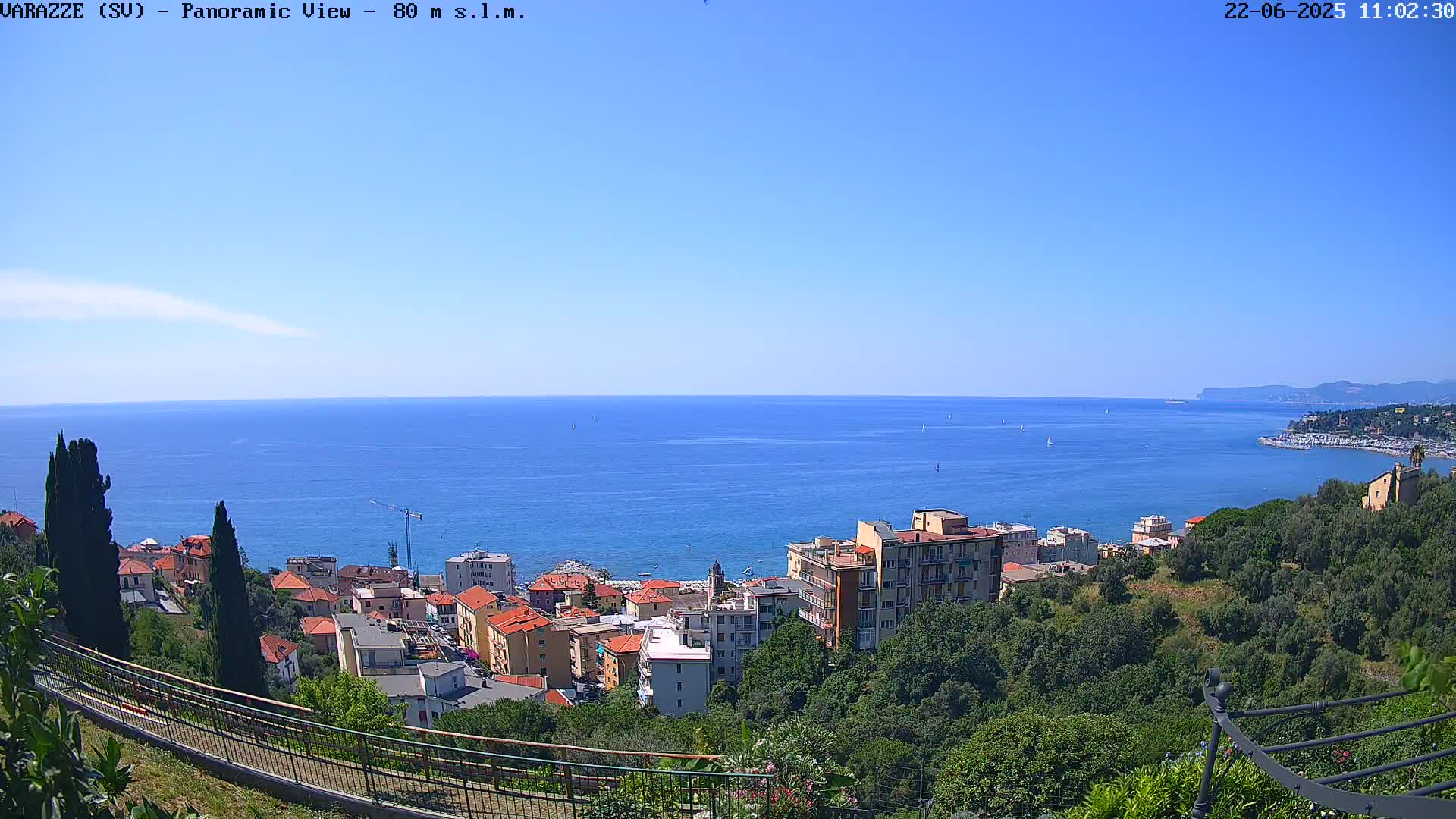 A coastal town with red-roofed buildings sits on a hillside overlooking a calm blue sea under a clear sky.